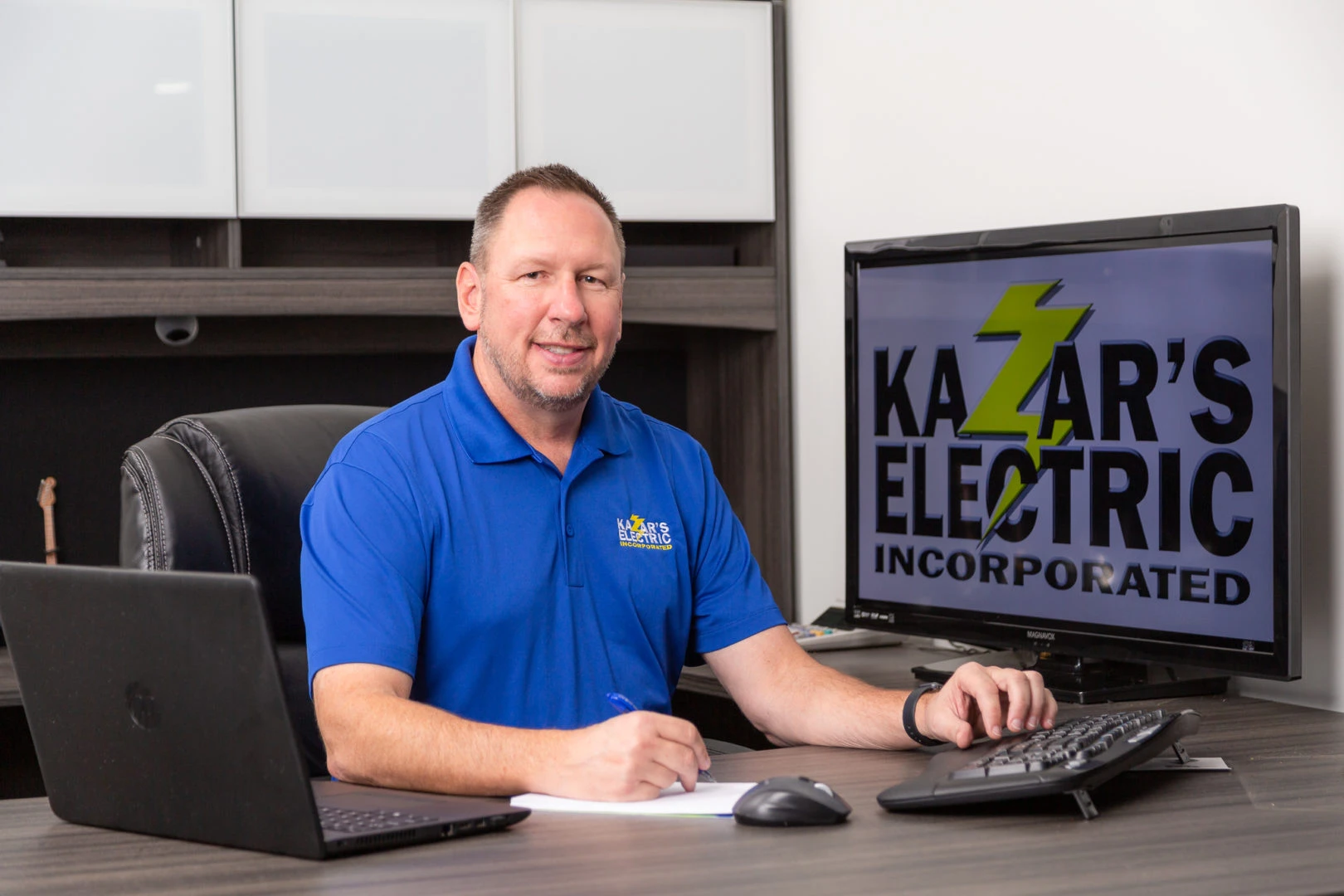 Man in blue polo at desk with Kazar's Electric branding, laptop, and monitor showing employment opportunities.