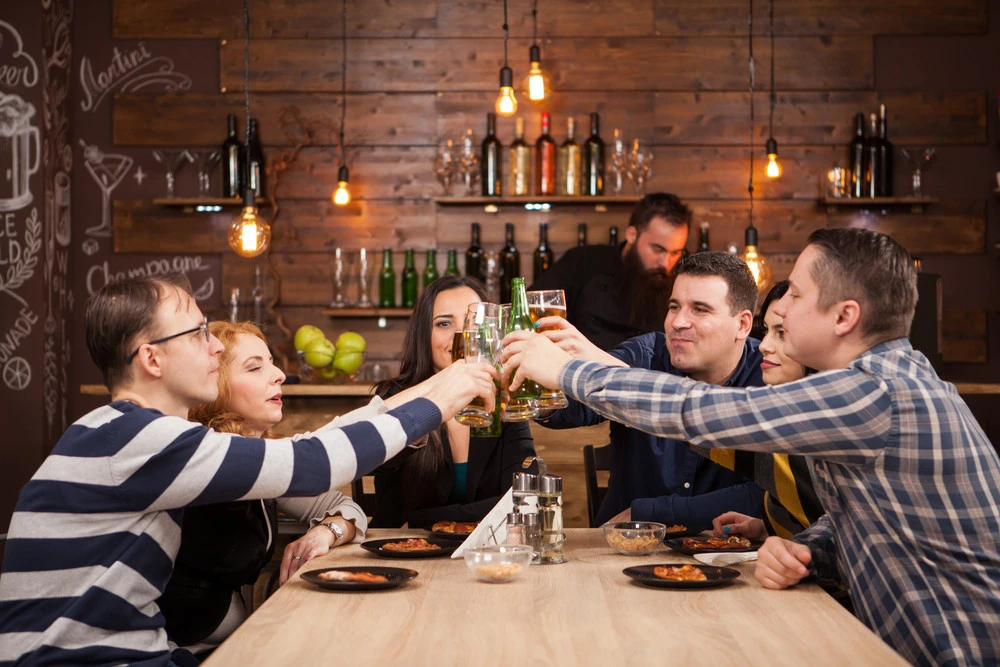 Group of friends toasting with drinks at a wooden table in a restaurant, highlighting social dining atmosphere and the importance of effective lighting for a vibrant dining experience.
