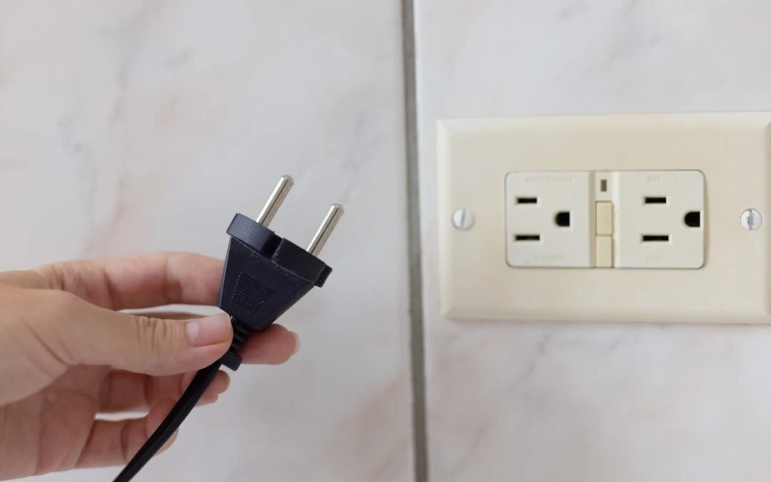 Hand holding a black electrical plug near a GFCI outlet on a wall, illustrating the importance of GFCI protection for home safety.