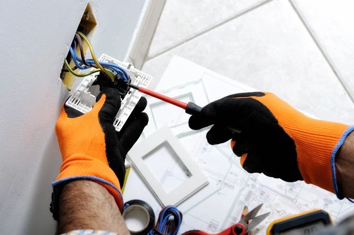 Electrician wearing safety gloves working on electrical wiring and outlet installation, emphasizing surge protection for electronics.